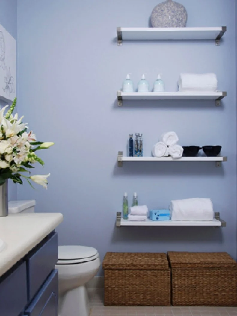 Blue bathroom with open floating shelves on wall with various bathroom items, wicker baskets, blue vanity cabinet, and clean design. 