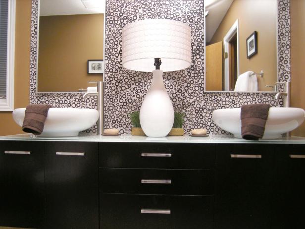 Dark vanity area with updated design, above counter sinks, lamp, dual mirrors, circular design wallpaper, and countertop decor. 