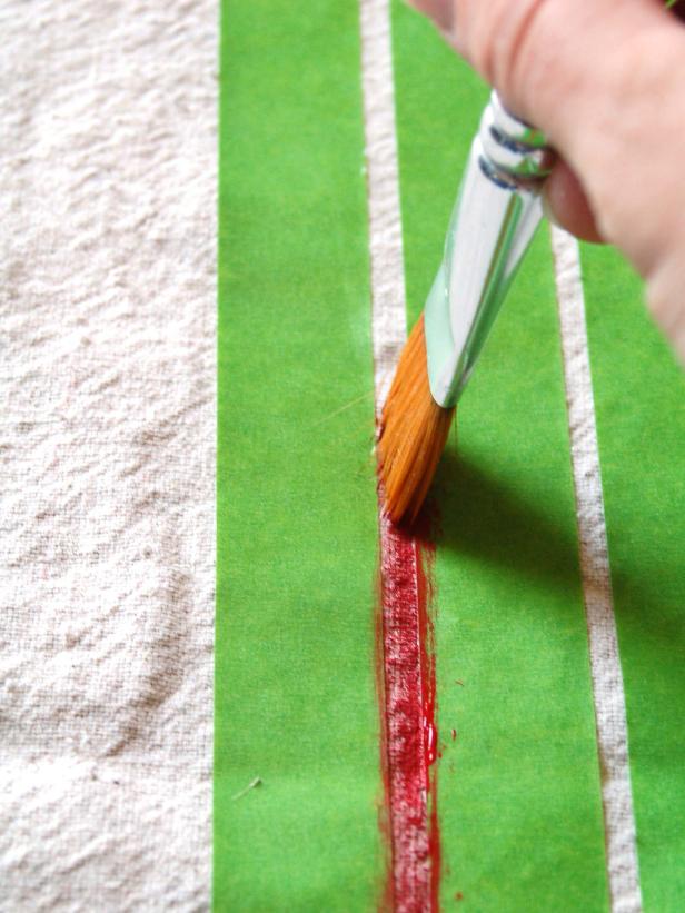Step 4A in creating grain sack barstool cushions is to select two seat pieces of fabric on which to paint the design. These will be the top sides of each cushion. Measuring side to side, find the center point and tape off two quarter-inch wide stripes. Using a one-inch brush and acrylic paint, paint the space between the taped lines. Allow some fabric to show through the paint for a time-worn look. Remove the tape.