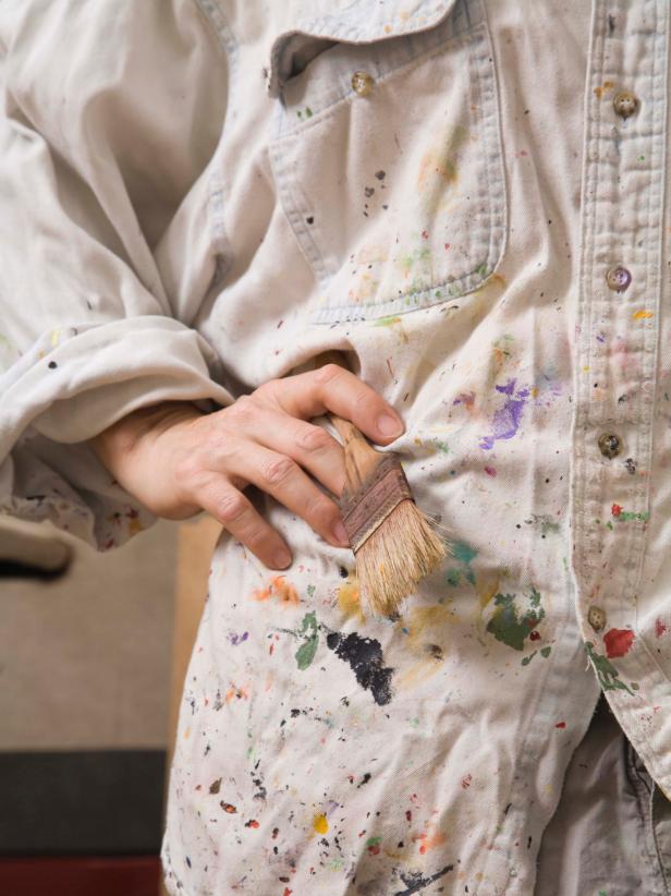 An artist stands in a smock covered with several colors of paint.