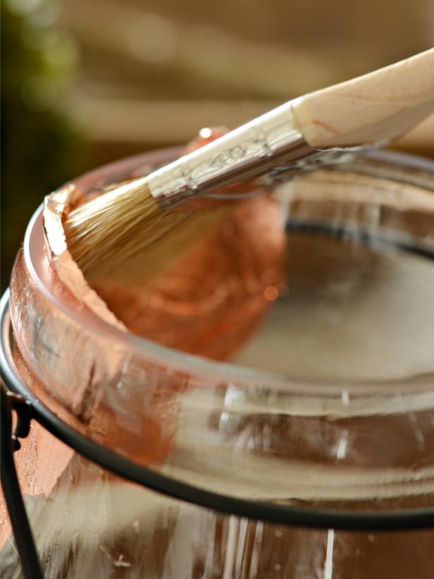 Working from top of the rim down, gently apply full sheets of copper leaf to entire interior of hurricane (Image 1). Smooth down with fingertip or chip brush (Image 2). Continue adding sheets until interior is completely covered. Buff copper with chip brush in a circular pattern to remove excess leaf then empty loosened bits from hurricane. The brush attachment on a vacuum is a great way to remove excess copper leaf. Insert candle or tealight and light as usual. Note: Never leave a burning candle unattended.