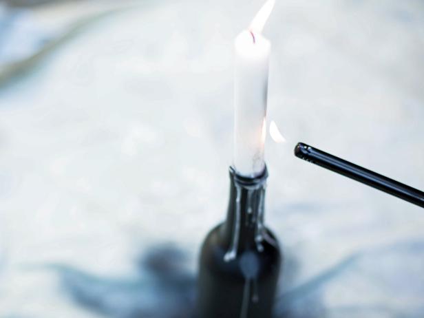 Tapered candles can easily lean then fall off the wine bottles and possibly break. To prevent this, place tapered candles into the openings at the top of each wine bottle, then light wicks immediately. Wax will drip down to the top of the opening, creating a bond between the bottle and the candle and helping each candle stand securely upright. Heavy drips and buildup along the exterior of the bottle adds drama to the overall look. Allow wax to accumulate all the way around the bottle before placing together as a group near your front porch or entryway.