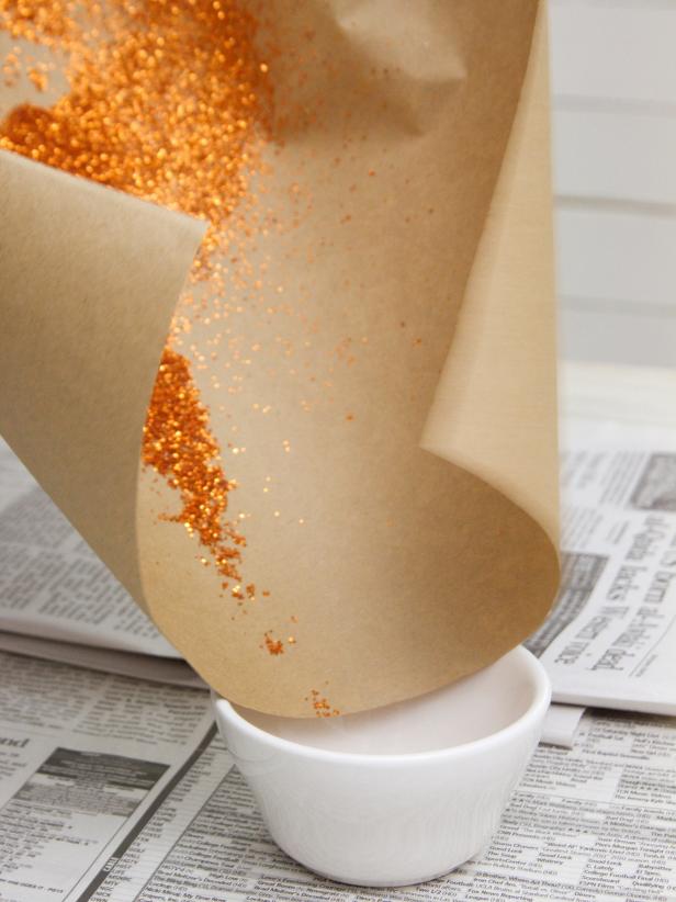 Pick up the pumpkin and shake off any glitter that didn't fully adhere. Set the pumpkin aside and fold the craft paper to funnel the excess glitter back into its original container. After removing glittery pumpkin from paper, carefully fold sides of paper into funnel shape and dump excess glitter into cup for later use.