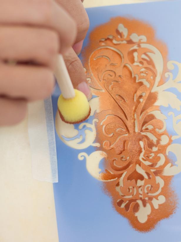 Remove the upper piece of masking tape and use it to secure the left side of the stencil to the pumpkin. Once it's in place, remove the lower piece of masking tape so the left side of the stencil is now lying flat against the pumpkin. Load up your brush and paint the left side of the stencil, using your fingers to firmly hold the stencil in place where necessary. Once that has dried, use a piece of masking tape to secure the right side of the stencil to the pumpkin, then remove the piece of tape of the left. Fill in the right portion of the stencil with craft paint, pressing down where necessary, and allow to dry before removing stencil.