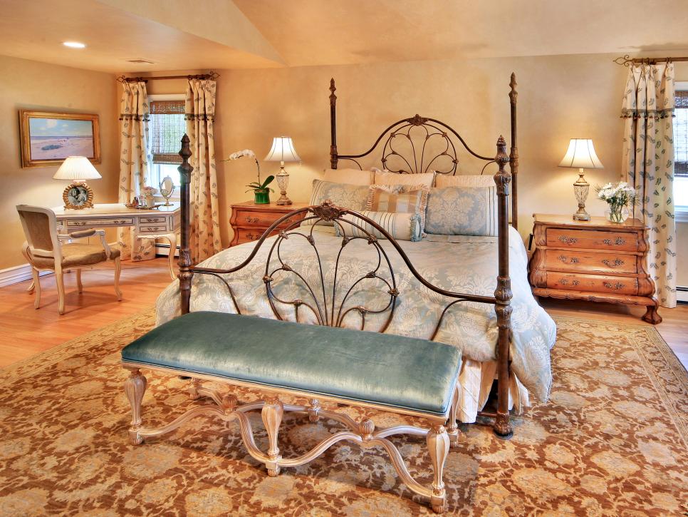 French Country bedroom with floral carpet, velvet bench and large iron bed frame.