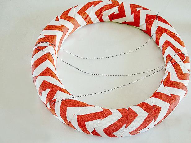 To start, wrap the orange fabric over the wreath form, slightly overlapping on the edges. Use your hot glue gun to attach the baker's twine onto the wreath, crisscrossing three times.