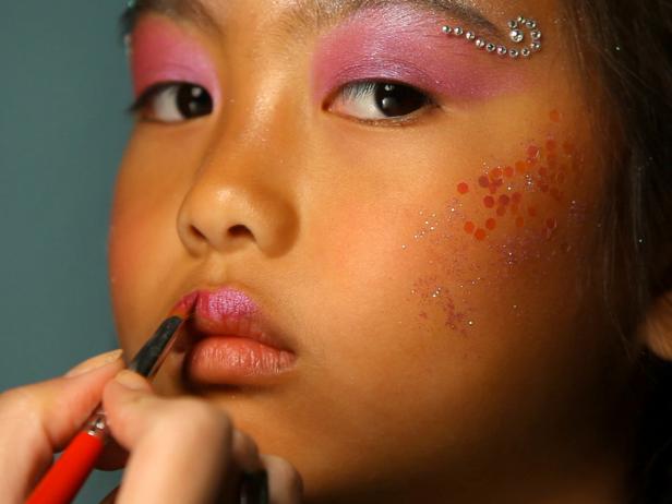 To apply fairy princess makeup, begin with a pearly eye shadow. Then add pink blush on the cheeks. Next, apply self-adhesive glittering jewels on forehead like a princess' tiara, and then apply additional jewels under eyebrows, on cheekbones or anywhere you like. Tip: You can find self-adhesive jewels in the scrapbooking section of your local craft store. Make sure your child doesn't have any adhesive allergies before continuing with this step. Keeping away from the sensitive eye area, dab face lotion on the cheeks and surrounding the jewel tiara on forehead. Have model close her eyes then apply cosmetic glitter where lotion was applied. Paint lips with a sparkly pink lipstick or tinted lip-gloss. Finish the look with a fairy princess costume.