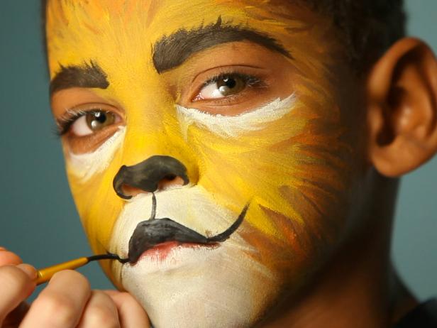 To give your child a ferocious lion’s face for Halloween, simply paint a basic orange and yellow face. Then paint their top lip black and extend curving line up cheeks slightly. Paint their bottom lip black, then add black dots to white area above top lip. Add whiskers by brushing white paint in small, short strokes around mouth and cheek area.