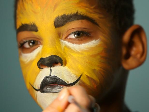 To give your child a ferocious lion’s face for Halloween, simply paint a basic orange and yellow face. Then paint their top lip black and extend curving line up cheeks slightly. Paint their bottom lip black, then add black dots to white area above top lip. Add whiskers by brushing white paint in small, short strokes around mouth and cheek area. Add a line of black paint under eyes, angling down the nose to create those striking big-cat eyes.