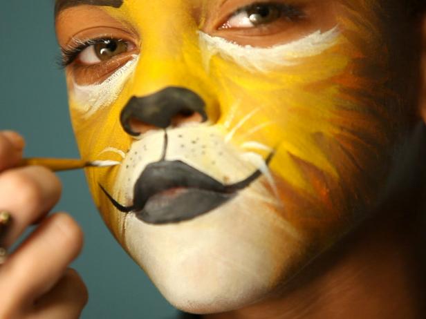 To give your child a ferocious lion’s face for Halloween, simply paint a basic orange and yellow face. Then paint their top lip black and extend curving line up cheeks slightly. Paint their bottom lip black, then add black dots to white area above top lip. Add whiskers by brushing white paint in small, short strokes around mouth and cheek area.