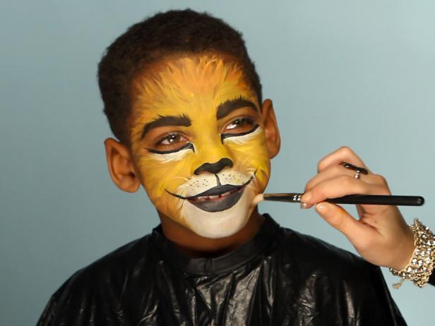 To give your child a ferocious lion’s face for Halloween, simply paint a basic orange and yellow face. Then paint their top lip black and extend curving line up cheeks slightly. Paint their bottom lip black, then add black dots to white area above top lip. Add whiskers by brushing white paint in small, short strokes around mouth and cheek area. Add a line of black paint under eyes, angling down the nose to create those striking big-cat eyes. Use a blending brush and brown eye shadow to shade down sides of nose, down the smile crease and chin, and create some brush texture around edges of face to look like fur. Add a few strokes of white to the forehead and chin for added depth and definition.