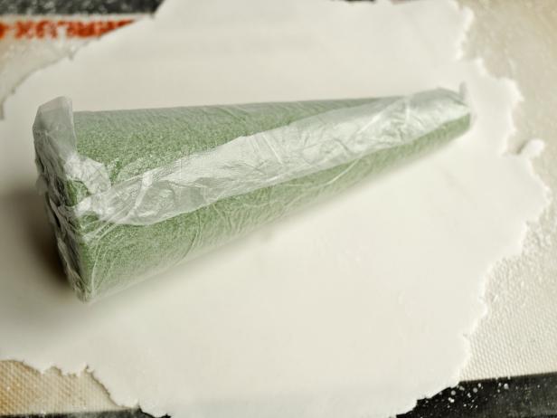 To make a Halloween candy topiary, first cover a foam cone in fondant. To make foam cones food-safe, cover them entirely with plastic wrap. Any large folds or wrinkles will show through the layer of fondant, so try to make this layer as smooth as possible. Note: Fondant dries out quickly, so cover with a damp paper towel to keep moist prior to rolling it out. Sprinkle rolling mat or clean work surface with powdered sugar to prevent fondant from sticking. Use a rolling pin to roll fondant to a 1/4-inch-thick piece large enough to cover one foam cone. Roll cone in fondant and trim away excess at top, seam and bottom with a knife. Fondant should overlap slightly at seam. Press edges together gently with fingers and neatly fold over top. Fondant will mostly be covered by candy, so it's okay if it's not perfectly smooth. Tip: Fondant can be colored with food coloring to create a different base color, if you'd like. Next add icing from piping bag where you want to place your candy pieces and cover in any pattern you like.