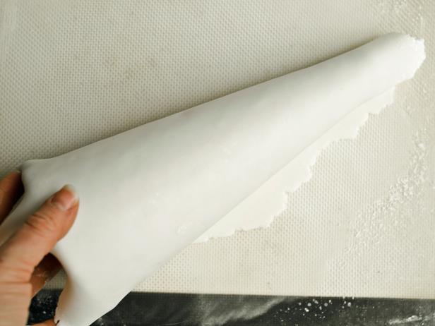To make a Halloween candy topiary, first cover a foam cone in fondant. To make foam cones food-safe, cover them entirely with plastic wrap. Any large folds or wrinkles will show through the layer of fondant, so try to make this layer as smooth as possible. Note: Fondant dries out quickly, so cover with a damp paper towel to keep moist prior to rolling it out. Sprinkle rolling mat or clean work surface with powdered sugar to prevent fondant from sticking. Use a rolling pin to roll fondant to a 1/4-inch-thick piece large enough to cover one foam cone. Roll cone in fondant and trim away excess at top, seam and bottom with a knife. Fondant should overlap slightly at seam. Press edges together gently with fingers and neatly fold over top. Fondant will mostly be covered by candy, so it's okay if it's not perfectly smooth. Tip: Fondant can be colored with food coloring to create a different base color, if you'd like. Next add icing from piping bag where you want to place your candy pieces and cover in any pattern you like.