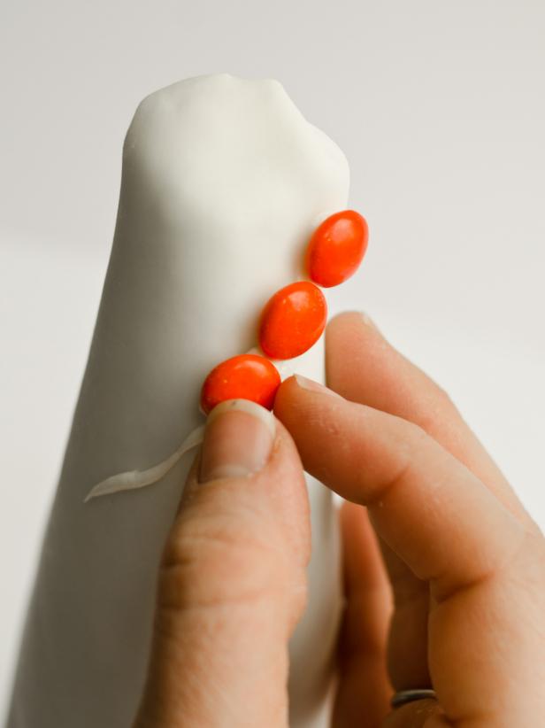 To make a Halloween candy topiary, first cover a foam cone in fondant. Then put icing in a piping bag fitted with a tip. Apply icing to a small section of fondant in desired design. Stick candy pieces immediately to icing. Continue covering cone until you're happy with the design. The design options are limitless: Use black licorice strands to create a spider web, write out messages or create graphic shapes, or give small black candies "legs" with black icing to make creepy, edible spiders. Food-safe markers can even be used to sketch a design on the fondant. Tip: Make candy topiaries only a couple of hours before a party, so the icing doesn't dry out too much. With the exception of the foam cone and plastic wrap, these topiaries are edible so you can allow kids to devour them as the party winds down.