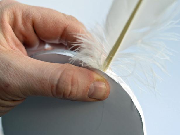 After applying a dab of hot glue to the feather's end and inserting it in the cut slit, Step 4E in creating feathered angel wings is to squeeze the sides of the foam core base together with your fingers until the glue cools. Repeat the whole process with all the feathers until the desired wing shape is created.