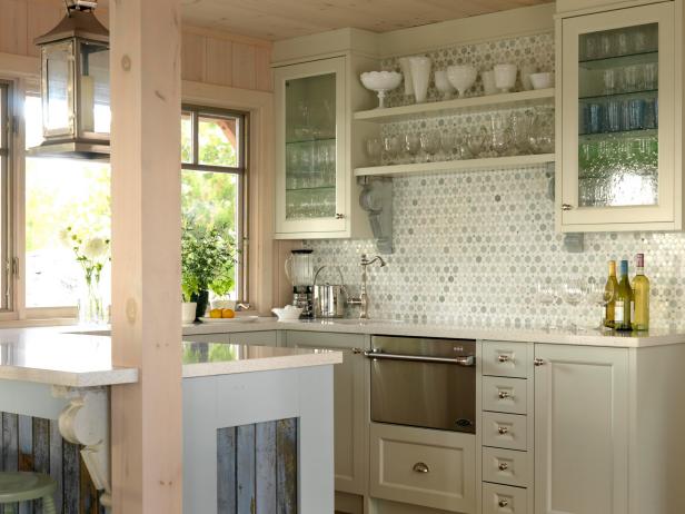 Two long runs of counter space give plenty of room in this cottage kitchen. Blue-painted antique corbels hold open shelving and support the peninsula overhang, bringing vintage-inspired whimsy to the space. Sarah chose three different cabinet colors to create a dynamic color palette. She used natural materials and a lighthearted color for the backsplash, and the seeded glass cabinet doors bring an extra touch of softness.