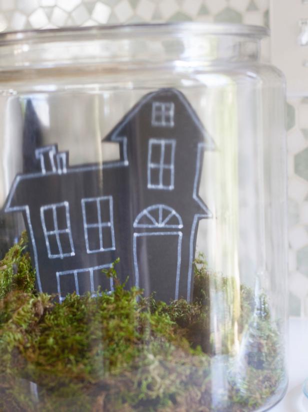 To create a spooky Halloween terrarium, add a 3-4" layer of sheet moss to the bottom of a large clear, lidded jar. Measure the height and width of the inside of the jar. Based on the dimensions, cut the shape of a haunted house out of a piece of black tag board, and then use a white paint pen to outline the windows and doors. Insert house into the moss and use clear tape to secure it to the back of the jar. Insert twigs into the moss to act as spooky, leafless trees.