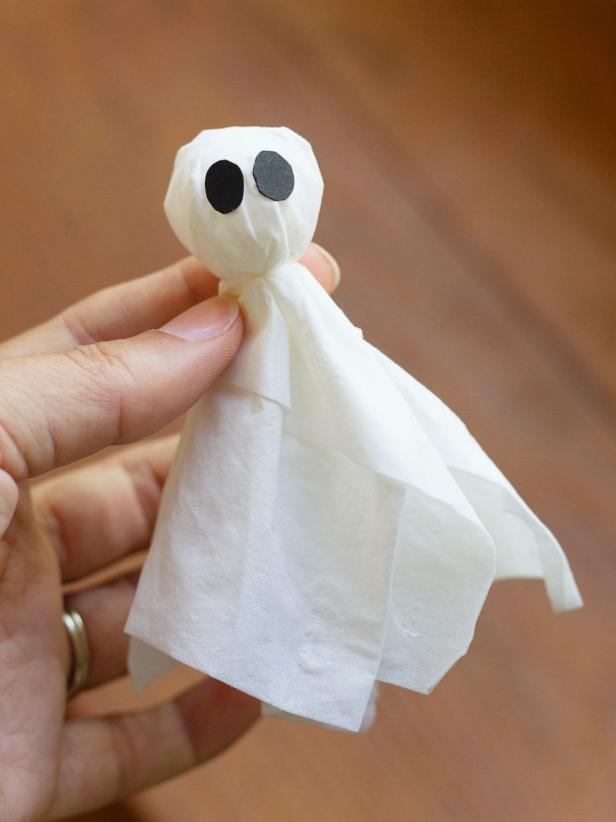 To create a spooky ghost to add to a Halloween-inspired terrarium, tear a tissue in half and layer the pieces to form a cross, then place a 1&quot; foam ball in the center. Gather the tissue around the foam ball and use fishing line to create the ghost's neck by tying a knot around the base of the foam ball. Cut out two black tag board ovals to act as eyes and attach them with glue. Shorten the prongs of a U-shaped floral pin with wire snips, and then push it into the top of the ghost's head. Knot one end of a piece of fishing line around the floral pin and attach the other end to the underside of the jar's lid with clear tape.