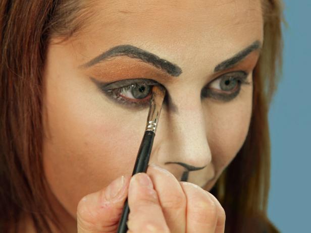 To turn yourself into an adorable cat for Halloween, cover your face with a blend of dark and light foundations to create the cat-face shape. Darken brows and create a bold cat eye with black eyeliner. Then color tip of nose black and add line from the nose to upper lip. Color top lip black. Apply a black shadow to the black areas for a more extreme cat eye. Use a white cream pencil and draw a small triangle just inside the black of the inner point. Using a large makeup brush, apply white shimmery eye shadow to the light highlights all around the face. Then, use black liquid eyeliner to accentuate the eyes by following the existing lines. Use the liner to dot on about five whisker spots above the lips on each side and draw three whiskers going out beside the nose. Add fun false eyelashes, cat ears, a tail and a collar to complete the look.