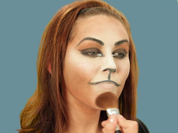 To turn yourself into an adorable cat for Halloween, cover your face with a blend of dark and light foundations to create the cat-face shape. Darken brows and create a bold cat eye with black eyeliner. Then color tip of nose black and add line from the nose to upper lip. Color top lip black. Apply a black shadow to the black areas for a more extreme cat eye. Use a white cream pencil and draw a small triangle just inside the black of the inner point. Using a large makeup brush, apply white shimmery eye shadow to the light highlights all around the face. Then, use black liquid eyeliner to accentuate the eyes by following the existing lines. Use the liner to dot on about five whisker spots above the lips on each side and draw three whiskers going out beside the nose. Add fun false eyelashes, cat ears, a tail and a collar to complete the look.