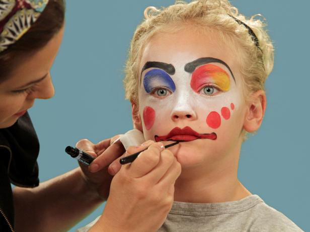 In order to give your child the perfect clown makeup for Halloween, use a makeup sponge and brush to apply white makeup to just the eyes and nose. Feather the makeup around the outside edges of the face so it fades softly. Use a makeup brush and blue face paint to draw a half circle over one eye following the brow line. Blend the blue and white. Do the same thing to half of the other eye, using red face paint. Next, add yellow face paint to the rest of the eyebrow and the upper lid. Blend slightly. Use a black eyeliner pencil to make a half circle above the child's natural eyebrow, then a line that follows the natural brow. Use a craft brush with red paint to make three small circles on one cheek. On the opposite cheek, draw a large circle with pink face paint. To make a clown mouth, first use red lip liner to line just outside the lips, then fill in. Extend the lines out from the edge of the natural lips and add little circles at the end. Outline lips with black liquid eyeliner. As a final accent, draw some lines just below the lower eyelid. Naturally curly hair (or a wig) is ready to clown around and just needs a nose. Oversized clothes or a clown costume finishes this big-top look.