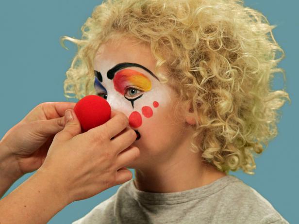 In order to give your child the perfect clown makeup for Halloween, use a makeup sponge and brush to apply white makeup to just the eyes and nose. Feather the makeup around the outside edges of the face so it fades softly. Use a makeup brush and blue face paint to draw a half circle over one eye following the brow line. Blend the blue and white. Do the same thing to half of the other eye, using red face paint. Next, add yellow face paint to the rest of the eyebrow and the upper lid. Blend slightly. Use a black eyeliner pencil to make a half circle above the child's natural eyebrow, then a line that follows the natural brow. Use a craft brush with red paint to make three small circles on one cheek. On the opposite cheek, draw a large circle with pink face paint. To make a clown mouth, first use red lip liner to line just outside the lips, then fill in. Extend the lines out from the edge of the natural lips and add little circles at the end. Outline lips with black liquid eyeliner. As a final accent, draw some lines just below the lower eyelid. Naturally curly hair (or a wig) is ready to clown around and just needs a nose. Oversized clothes or a clown costume finishes this big-top look.
