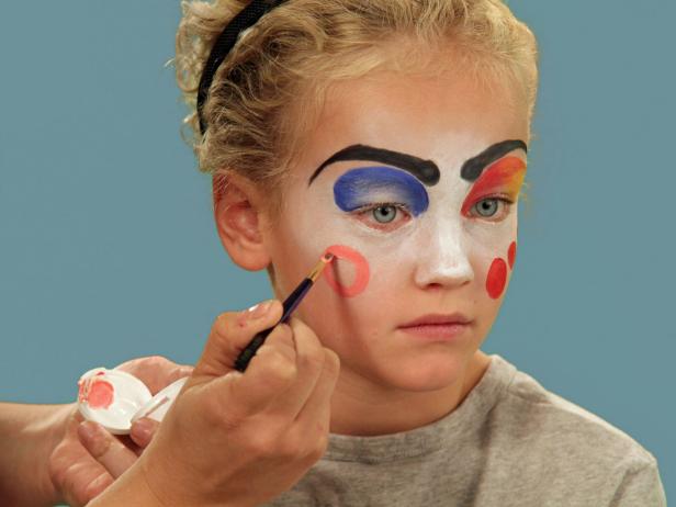 In order to give your child the perfect clown makeup for Halloween, use a makeup sponge and brush to apply white makeup to just the eyes and nose. Feather the makeup around the outside edges of the face so it fades softly. Use a makeup brush and blue face paint to draw a half circle over one eye following the brow line. Blend the blue and white. Do the same thing to half of the other eye, using red face paint. Next, add yellow face paint to the rest of the eyebrow and the upper lid. Blend slightly. Use a black eyeliner pencil to make a half circle above the child's natural eyebrow, then a line that follows the natural brow. Use a craft brush with red paint to make three small circles on one cheek. On the opposite cheek, draw a large circle with pink face paint. To make a clown mouth, first use red lip liner to line just outside the lips, then fill in. Extend the lines out from the edge of the natural lips and add little circles at the end. Outline lips with black liquid eyeliner. As a final accent, draw some lines just below the lower eyelid. Naturally curly hair (or a wig) is ready to clown around and just needs a nose. Oversized clothes or a clown costume finishes this big-top look.