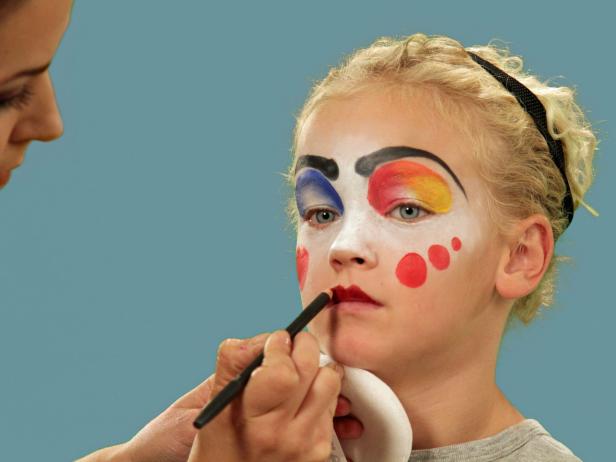 In order to give your child the perfect clown makeup for Halloween, use a makeup sponge and brush to apply white makeup to just the eyes and nose. Feather the makeup around the outside edges of the face so it fades softly. Use a makeup brush and blue face paint to draw a half circle over one eye following the brow line. Blend the blue and white. Do the same thing to half of the other eye, using red face paint. Next, add yellow face paint to the rest of the eyebrow and the upper lid. Blend slightly. Use a black eyeliner pencil to make a half circle above the child's natural eyebrow, then a line that follows the natural brow. Use a craft brush with red paint to make three small circles on one cheek. On the opposite cheek, draw a large circle with pink face paint. To make a clown mouth, first use red lip liner to line just outside the lips, then fill in. Extend the lines out from the edge of the natural lips and add little circles at the end. Outline lips with black liquid eyeliner. As a final accent, draw some lines just below the lower eyelid. Naturally curly hair (or a wig) is ready to clown around and just needs a nose. Oversized clothes or a clown costume finishes this big-top look.