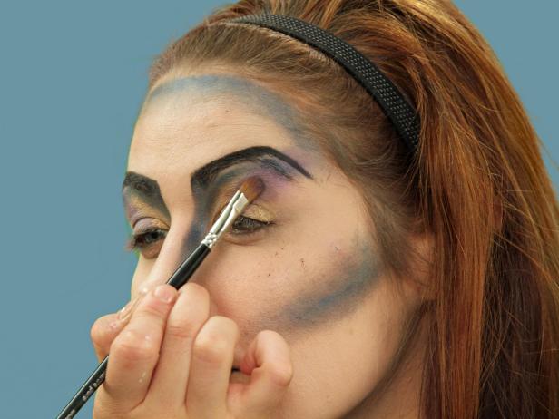 With an eye shadow brush, add dark purple eye shadow to the outside of the eye lids, carrying the color just under the blue eye shadow. Apply the purple to the inside corner of the eye and blend down onto the nose. Grab a larger makeup brush and sweep the purple eye shadow over the center of the cheekbones.