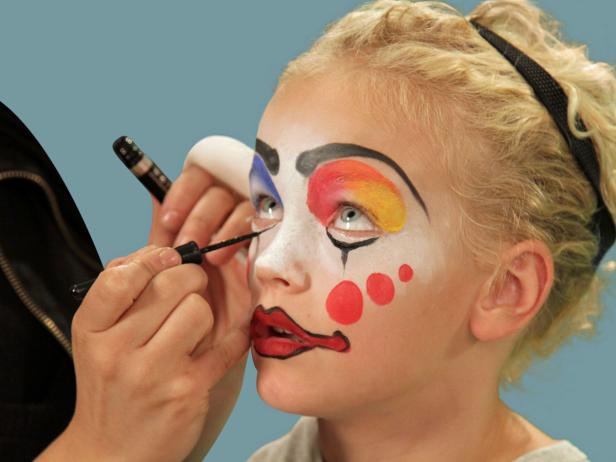 In order to give your child the perfect clown makeup for Halloween, use a makeup sponge and brush to apply white makeup to just the eyes and nose. Feather the makeup around the outside edges of the face so it fades softly. Use a makeup brush and blue face paint to draw a half circle over one eye following the brow line. Blend the blue and white. Do the same thing to half of the other eye, using red face paint. Next, add yellow face paint to the rest of the eyebrow and the upper lid. Blend slightly. Use a black eyeliner pencil to make a half circle above the child's natural eyebrow, then a line that follows the natural brow. Use a craft brush with red paint to make three small circles on one cheek. On the opposite cheek, draw a large circle with pink face paint. To make a clown mouth, first use red lip liner to line just outside the lips, then fill in. Extend the lines out from the edge of the natural lips and add little circles at the end. Outline lips with black liquid eyeliner. As a final accent, draw some lines just below the lower eyelid. Naturally curly hair (or a wig) is ready to clown around and just needs a nose. Oversized clothes or a clown costume finishes this big-top look.