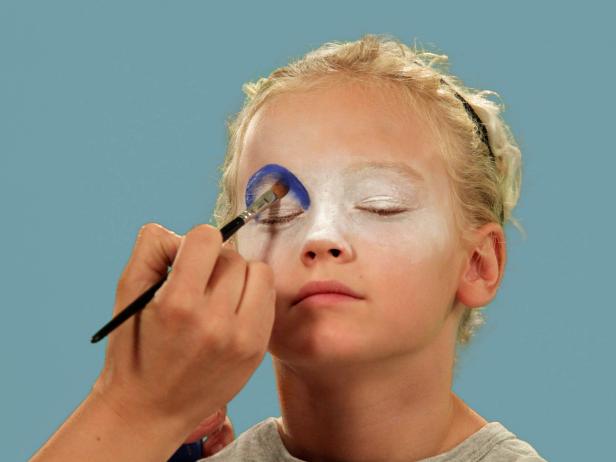 In order to give your child the perfect clown makeup for Halloween, use a makeup sponge and brush to apply white makeup to just the eyes and nose. Feather the makeup around the outside edges of the face so it fades softly. Use a makeup brush and blue face paint to draw a half circle over one eye following the brow line. Blend the blue and white. Do the same thing to half of the other eye, using red face paint. Next, add yellow face paint to the rest of the eyebrow and the upper lid. Blend slightly. Use a black eyeliner pencil to make a half circle above the child's natural eyebrow, then a line that follows the natural brow. Use a craft brush with red paint to make three small circles on one cheek. On the opposite cheek, draw a large circle with pink face paint. To make a clown mouth, first use red lip liner to line just outside the lips, then fill in. Extend the lines out from the edge of the natural lips and add little circles at the end. Outline lips with black liquid eyeliner. As a final accent, draw some lines just below the lower eyelid. Naturally curly hair (or a wig) is ready to clown around and just needs a nose. Oversized clothes or a clown costume finishes this big-top look.