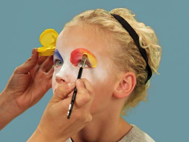 In order to give your child the perfect clown makeup for Halloween, use a makeup sponge and brush to apply white makeup to just the eyes and nose. Feather the makeup around the outside edges of the face so it fades softly. Use a makeup brush and blue face paint to draw a half circle over one eye following the brow line. Blend the blue and white. Do the same thing to half of the other eye, using red face paint. Next, add yellow face paint to the rest of the eyebrow and the upper lid. Blend slightly. Use a black eyeliner pencil to make a half circle above the child's natural eyebrow, then a line that follows the natural brow. Use a craft brush with red paint to make three small circles on one cheek. On the opposite cheek, draw a large circle with pink face paint. To make a clown mouth, first use red lip liner to line just outside the lips, then fill in. Extend the lines out from the edge of the natural lips and add little circles at the end. Outline lips with black liquid eyeliner. As a final accent, draw some lines just below the lower eyelid. Naturally curly hair (or a wig) is ready to clown around and just needs a nose. Oversized clothes or a clown costume finishes this big-top look.