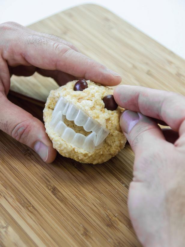 To make rice cereal monsters, melt 8 tablespoons of unsalted butter and 12 cups mini marshmallows in a large pot over medium low heat until all marshmallows are melted. Remove from heat and stir in1/2 teaspoon salt, 1 teaspoon vanilla and 12 cups rice cereal until fully coated with marshmallow mixture. Pour mixture into a large greased bowl or baking sheet. Use a little butter to grease hands and form balls (about 65 grams each) by pressing firmly and rolling between palms. Place tightly pressed balls on a baking sheet and refrigerate for at least 1 hour before proceeding. Using a serrated knife, carefully cut a wedge in the chilled balls large enough for the plastic vampire teeth to fit inside. Save cut wedges for a later step. Place the teeth inside the opening of the cereal ball. Cut the ends from both sides of the wedge about 1/2 inch long. Insert the cut pieces into the opening on each side of the monster's mouth to close up the sides. Make holes for the eyes. Add candy pieces to the holes. Store in an airtight container until ready to serve.