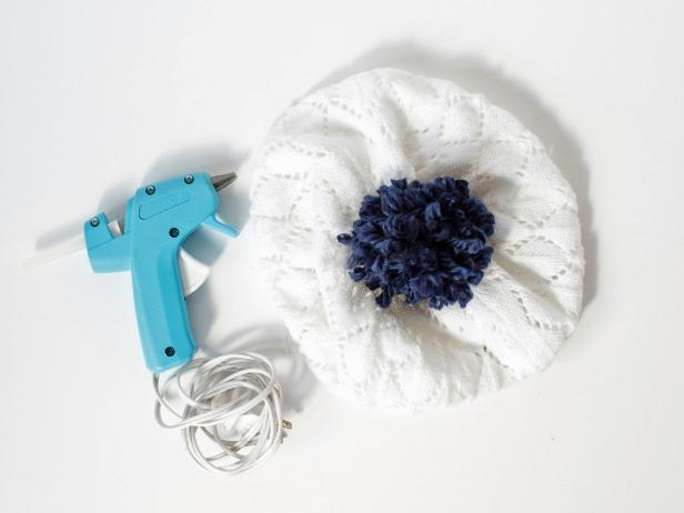 Using scissors, cut the two loop ends of the yarn so that they fluff out creating a yarn “poof” ball.Using a hot glue gun, secure this poof top the top of the knit hat.