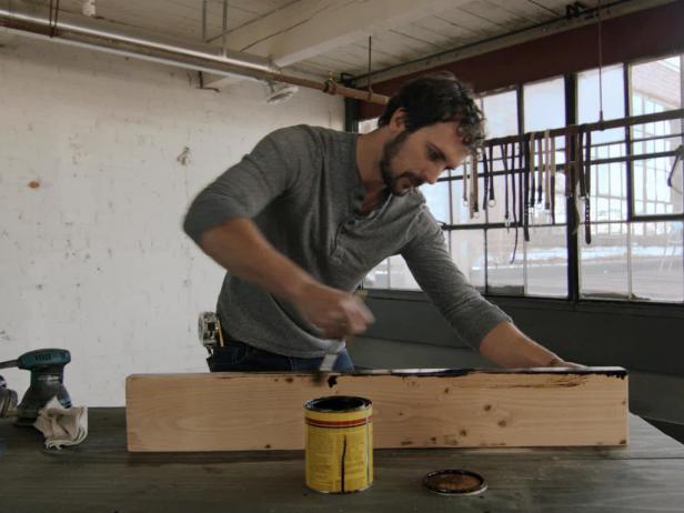 Use a paintbrush to apply a thick coat of stain to plank of wood to make a floating shelf and smooth it out with a rag to highlight the wood's natural grain.