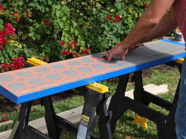 At this stage you’re ready to stain all your lumber if you haven’t already, including your ¾” dowels, the 1x2x6 and assembled bar top. Using a foam brush, apply stain, then wipe off the excess. Once dry, move on to the decorative paint process. We went with a Moroccan inspired design and a clever tracing trick! Place a can lid in one corner so it fits completely on the stained board. Using a ½” flat brush, trace around the lid with one of your paint colors- we used coral. Repeat, until entire bar top is filled with circular design. Once dry, repeat the process with a different color paint. To achieve the cool geometric effect, just reposition the starting point so that the lid overlaps four of the coral circles and begin tracing. Repeat until you have filled up the bar top. Once dry, top off with a coat of Polyurethane to protect your masterpiece!