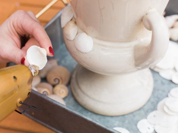 Start in one area of the urn and begin by adding a simple row of similar looking shells using the hot glue gun. You’re essentially creating bands up and down the urn- using one particular type of shell. Place a bead of hot glue onto the perimeter of the shell then push it into place on the urn. It helps to let the shape of your particular vessel and the shell dictate your design. At the base of the urn, leave a little space, so that the shell’s edge doesn’t chip off or scratch the surface of the table it’s sitting on and remember to rotate the shells in different directions, as well for visual interest and better coverage. We used shells in a variety of shapes and colors.