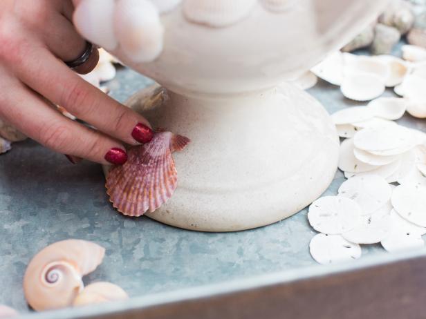 Start in one area of the urn and begin by adding a simple row of similar looking shells using the hot glue gun. You’re essentially creating bands up and down the urn- using one particular type of shell. Place a bead of hot glue onto the perimeter of the shell then push it into place on the urn. It helps to let the shape of your particular vessel and the shell dictate your design. At the base of the urn, leave a little space, so that the shell’s edge doesn’t chip off or scratch the surface of the table it’s sitting on and remember to rotate the shells in different directions, as well for visual interest and better coverage. We used shells in a variety of shapes and colors.