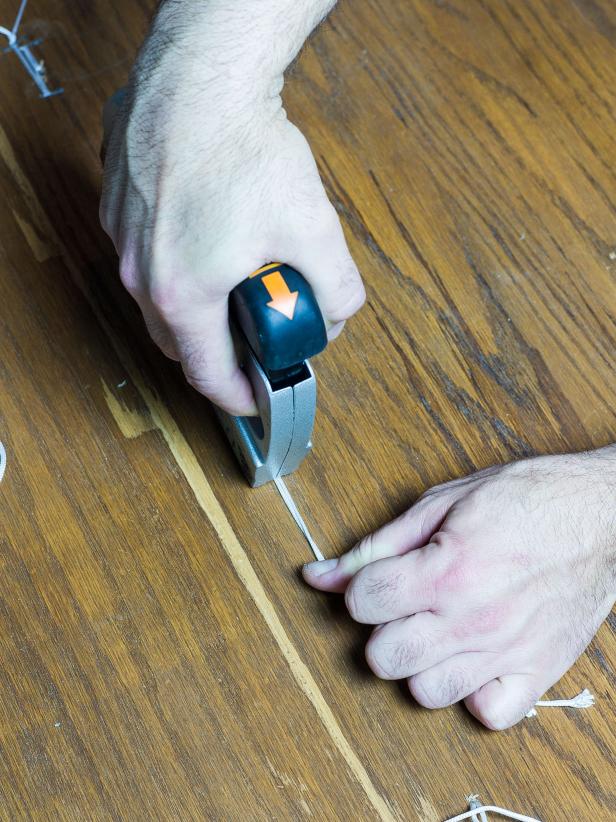 Pull the cording all the way through to the bottom of the ottoman top. Repeat for all buttons. When all buttons have been threaded, place the ottoman top face down on a flat surface. Pull each pair of cording ends very tightly in one direction and fasten to the bottom of the ottoman top with a staple. Then pull the cording in the opposite direction and staple again. For added security you can tie a knot with the two ends to make sure it doesnât slip through the staple. Cut away any excess.