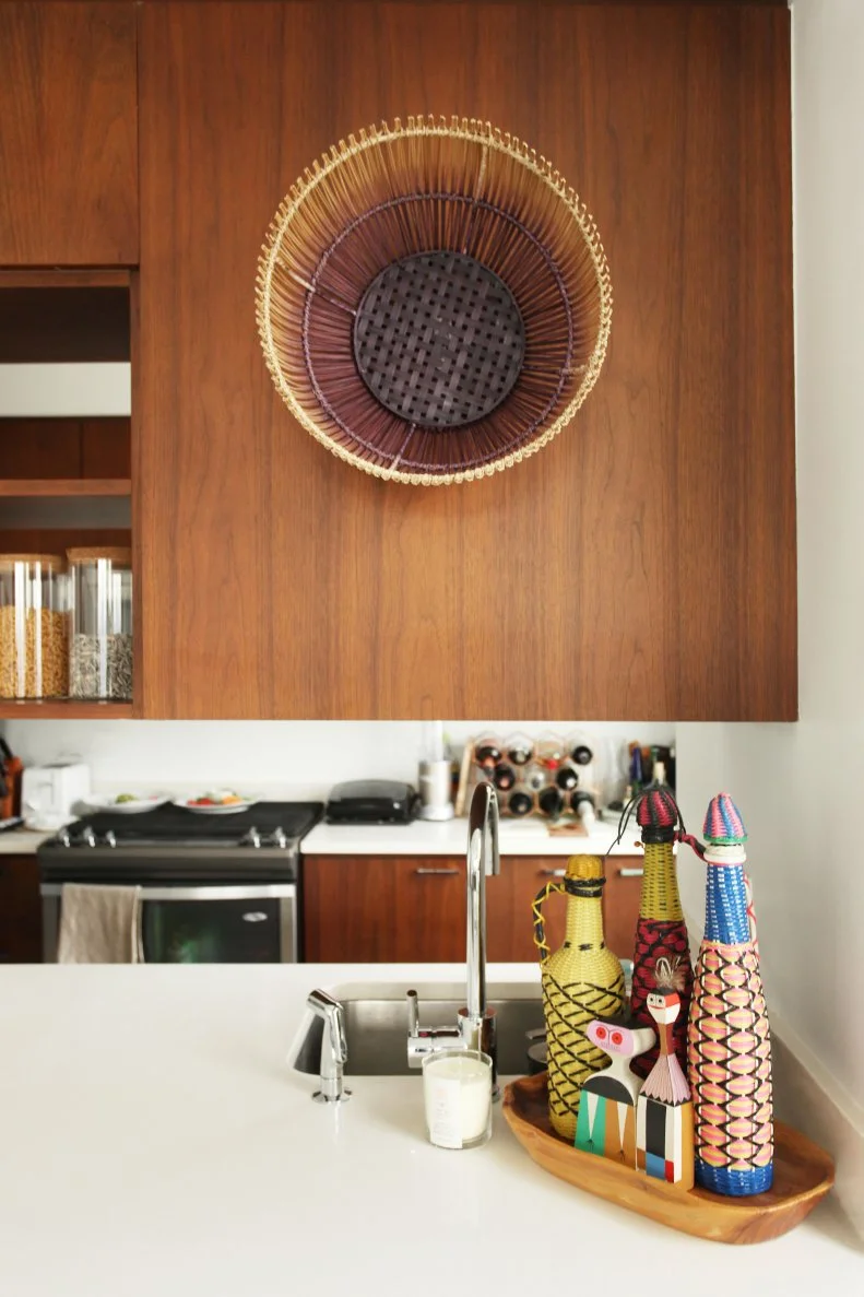 The kitchen features modern design complemented by warm, rustic wood elements. A large wicker basket finds new life here as an art installation giving depth as well as texture to the space. For color, an assortment of wine bottles handmade in South Africa add an element of global culture.

