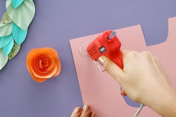 Make a flower by cutting a spiral into another piece of origami paper. Cut a wave into the outside edge of the spiral. Roll it up to get a flower shape. Put down a dollop or hot glue onto a piece of cardstock and press the flower down onto it. Once it dries, cut it out from the cardstock.