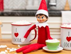 A toy elf on a table with marshmallows and a tiny hot cocoa mug