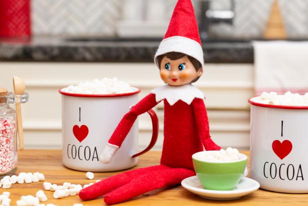 A toy elf on a table with marshmallows and a tiny hot cocoa mug