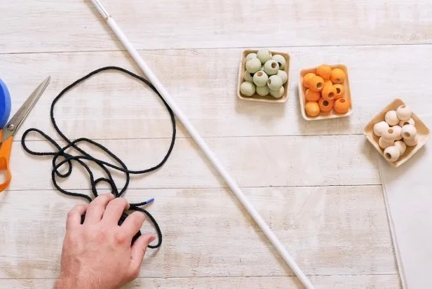 The Crafty Lumberjacks teach you how to make a funky, macrame beaded curtain with a plant holder in the middle. To start, you will need navy rope cording, wooden ball spacer beads, orange and teal acrylic paint, a tension rod, scissors, painter's tape, tape measure and a piece of sandpaper.