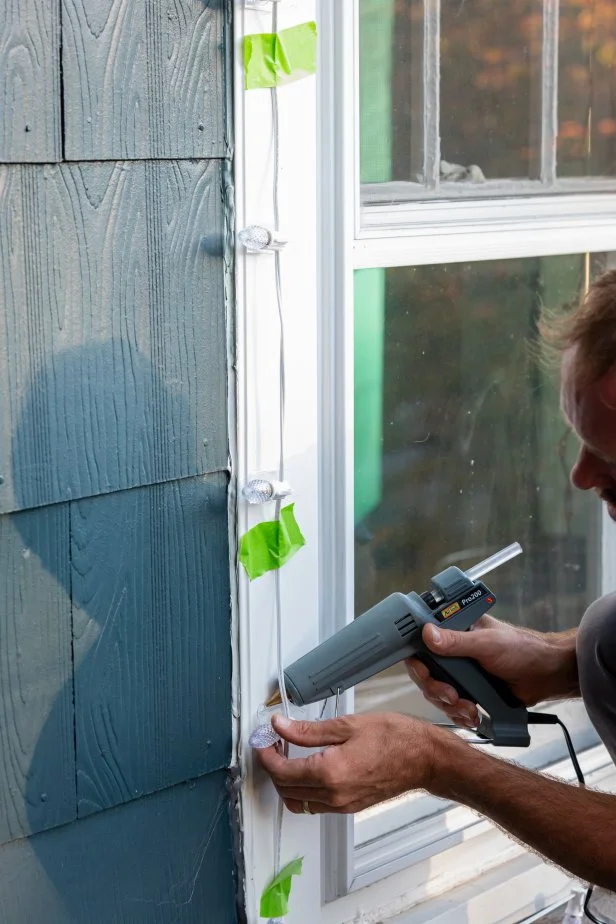 Hot glue is applied to a window sill to hold Christmas lights in place. Painter's tape is used as a temporary hold while the hot glue sets.