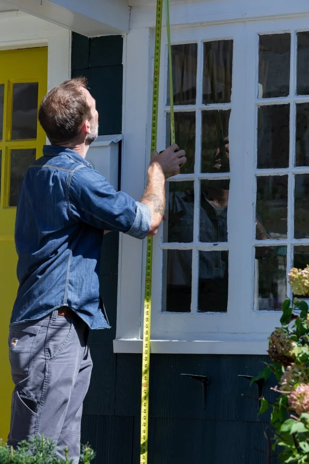 A measuring tape is used to measure a window on the exterior of a home for accurate fitting of Christmas string lights.