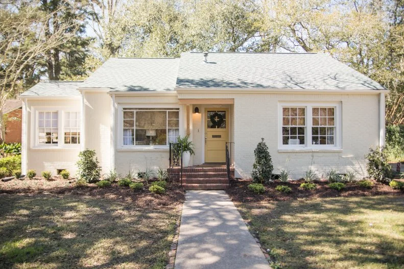 A cream exterior, yellow door, wood accent over the entryway, and green architectural roof have completely transformed the Douglas house