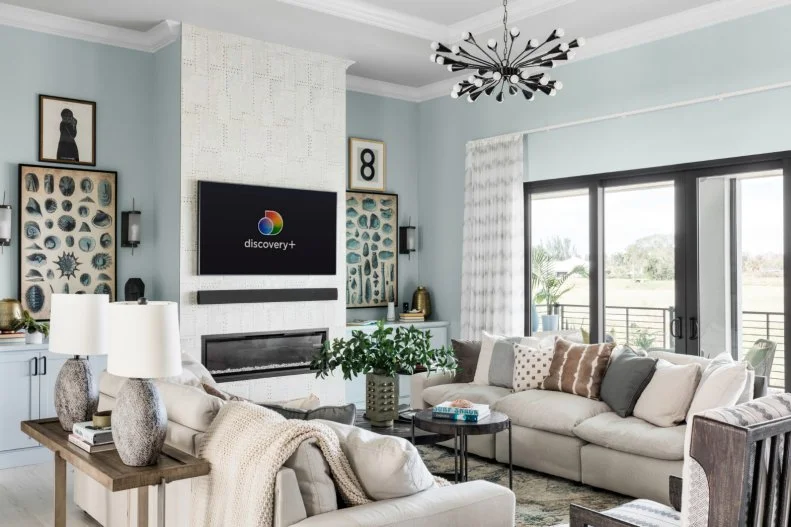 Soothing ocean tones bring a calm and tranquil feel to the living room at HGTV Smart Home 2021. Ample seating in front of the fireplace invites relaxation while natural light illuminates the room from the large glass doors that lead outside.