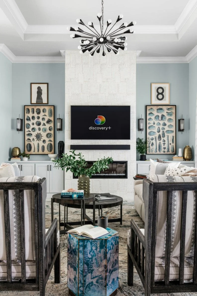 The vaulted ceiling uses multiple levels of crown molding to draw the eye upward to the industrial chandelier and enhance the room’s spacious feel. Designer Tiffany Brooks chose to encase the massive fireplace with a shell-like finish that adds a fresh, coastal feel to the living space.