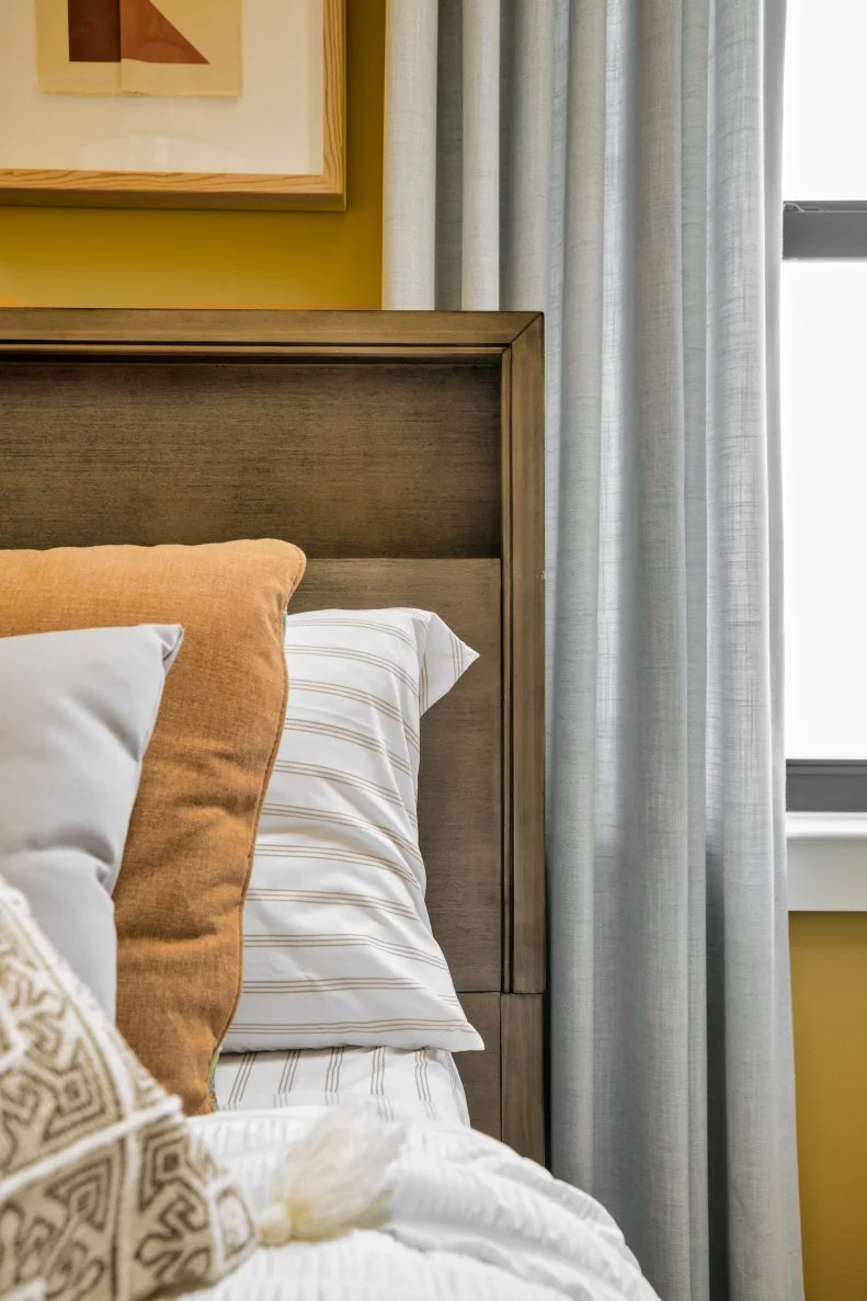 A handsome headboard in wood adds a natural texture to this warm and spacious guest suite. Soft stripes add a classic pattern to the bedding while tones of soft blue and warm orange play off the wall color and drapes to give the room a cohesive feel.